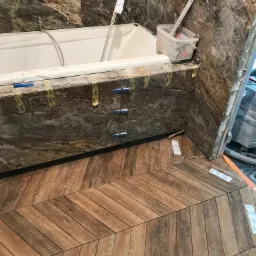 Montaż płytek imitujących drewno na podłodze i marmurowych płytek na ścianie w trakcie prac wykończeniowych łazienki. Widoczne narzędzia i materiały budowlane.