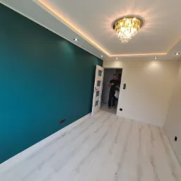 Świeżo pomalowane wnętrze z turkusową ścianą, białymi panelami podłogowymi i ozdobnym żyrandolem, widoczny mężczyzna w przejściu