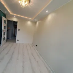 Jasne, odświeżone wnętrze z panelami podłogowymi, kremowymi ścianami, ozdobnym żyrandolem i podwieszanym sufitem z oświetleniem punktowym, widok na otwarte drzwi do kolejnego pomieszczenia...