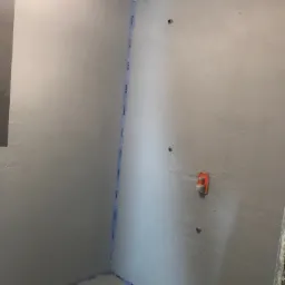 Świeżo położona szara gładź na ścianach w trakcie remontu łazienki, zabezpieczona niebieską taśmą Mapei wzdłuż krawędzi podłogi i ścian, widoczne puszki instalacyjne i wyciągnięta linka traserska.