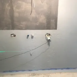 Ściana w trakcie przygotowania do dalszych prac, widoczne ślady po szpachlowaniu, otwory na instalacje elektryczne i poziomica laserowa wyznaczająca linię prostą.