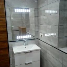 Szara łazienka z podświetlanym lustrem, białą szafką pod umywalką i drewnianą ścianą po lewej stronie.