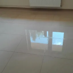 Jasne, kwadratowe płytki podłogowe z widocznym odbiciem okna i nieba, świeżo po ułożeniu, z grzejnikiem na ścianie w tle.