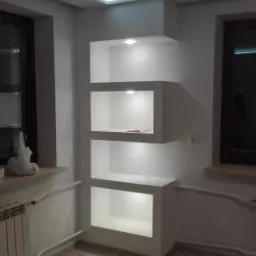 Białe, podświetlane p&oacute;łki wnękowe z płyty gipsowo-kartonowej, w nowocześnie wykończonym wnętrzu z drewnopodobną podłogą i oświetleniem LED na suficie.