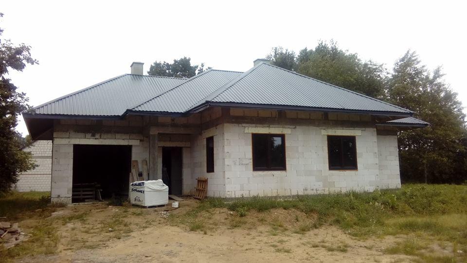 Niedokończony dom jednorodzinny z szarą blachodachówką i ścianami z białego bloczka, widoczne okna z ciemnymi ramami i materiały budowlane przed budynkiem.