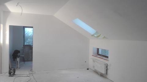 Poddasze po remoncie: białe ściany, okno dachowe z taśmą malarską, okno w ścianie, grzejnik zabezpieczony folią, wentylator i worek gipsu w tle.