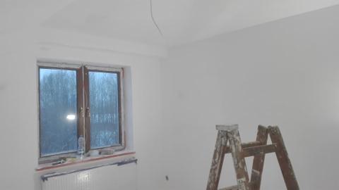 Świeżo pomalowane na biało wnętrze pokoju z drewnianym oknem, grzejnikiem i drabiną malarską w rogu.