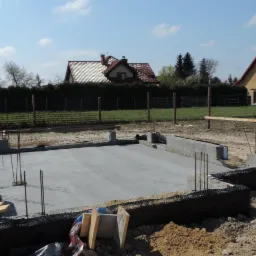 Wylany fundament z izolacją pionową i wystającymi prętami zbrojeniowymi, widok na działkę z dwoma domami w tle.
