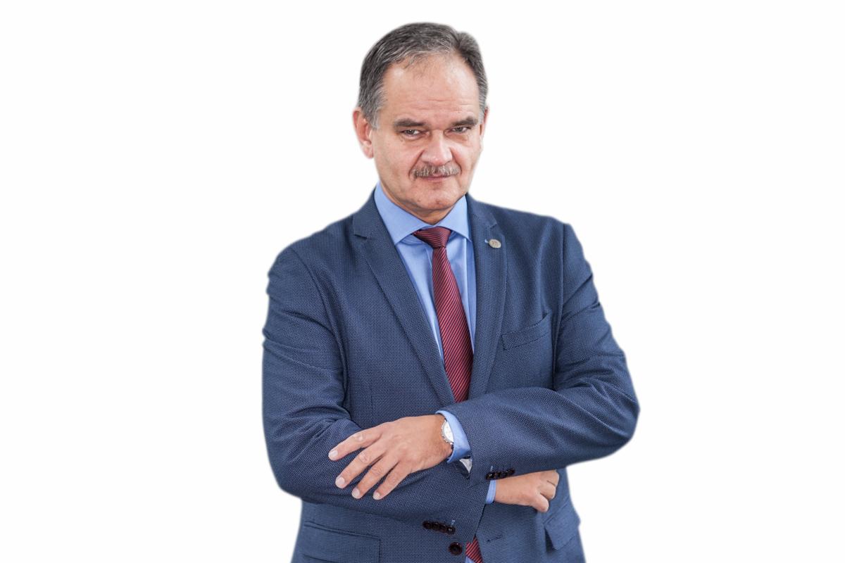 Portret mężczyzny w niebieskim garniturze i czerwonym krawacie, z założonymi rękami, uśmiechającego się do kamery na białym tle.