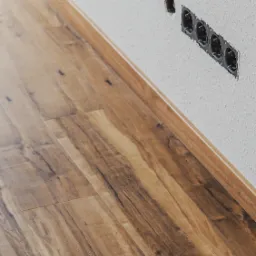 Ułożone panele podłogowe w odcieniach brązu, widoczne listwy przypodłogowe i puszka elektryczna bez osprzętu na białej ścianie.