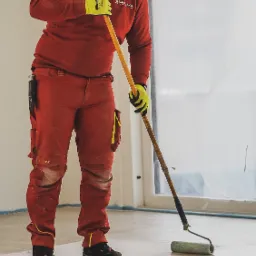 Mężczyzna w czerwonym kombinezonie i żółtych rękawicach rozprowadza masę szpachlową na podłodze za pomocą wałka na długim kiju, okno zabezpieczone folią malarską w tle.