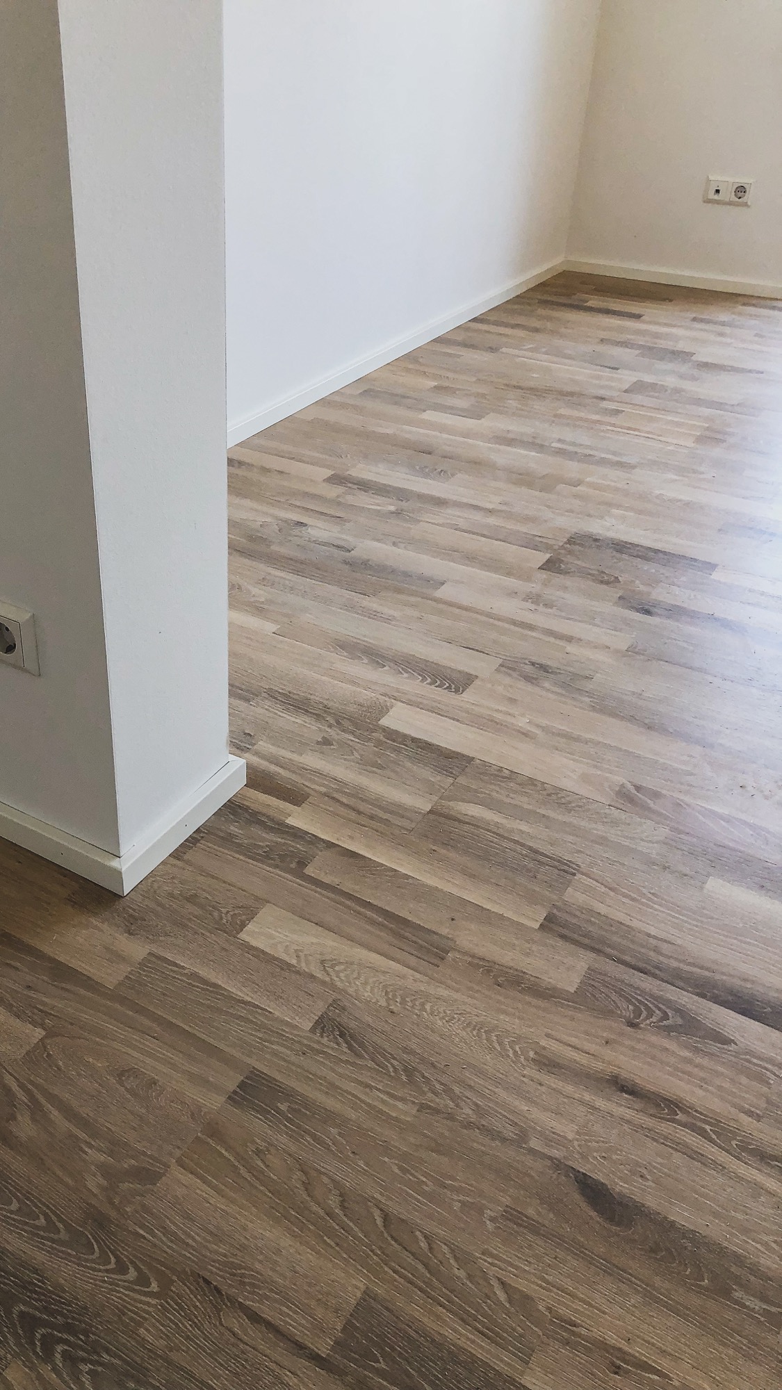 Nowo ułożone panele podłogowe w odcieniach brązu i beżu, widoczne gniazdka elektryczne na białej ścianie i biała listwa przypodłogowa.