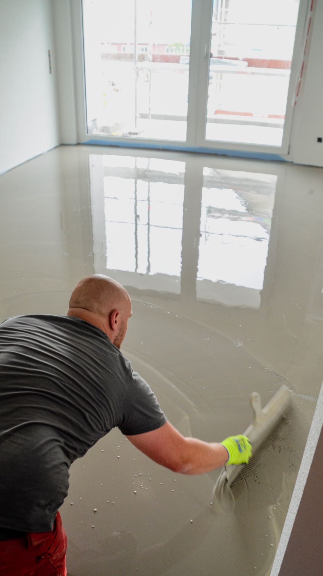 Pracownik w neonowych rękawicach rozprowadza masę szpachlową na podłodze w jasnym pomieszczeniu z dużym oknem, widoczne refleksy światła.