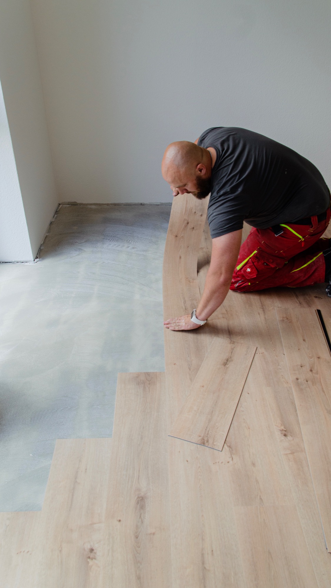 Mężczyzna w czerwonych spodniach montuje jasne panele podłogowe w pomieszczeniu z przygotowanym podłożem.