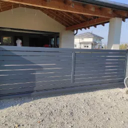 Nowoczesna, przesuwna brama wjazdowa z poziomych, szarych paneli, zamontowana przed domem jednorodzinnym z widocznym podjazdem wysypanym żwirem i elementami dekoracyjnego oświetlenia zewnętrznego.