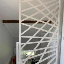 Biała, ażurowa ścianka działowa z motywem geometrycznym, oddzielająca schody od reszty pomieszczenia, z widocznymi elementami drewnianymi stropu.