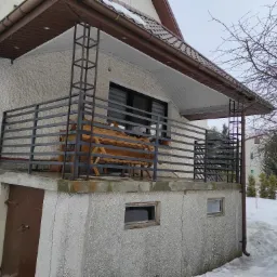 Balkon z ciemną metalową konstrukcją, drewnianym siedziskiem i widokiem na zasypany śniegiem ogród w pochmurny dzień.