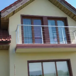 Balkon z nowoczesną balustradą ze stali nierdzewnej z cienkimi poziomymi linkami, zamontowaną na budynku z beżową elewacją i brązowymi ramami okiennymi.