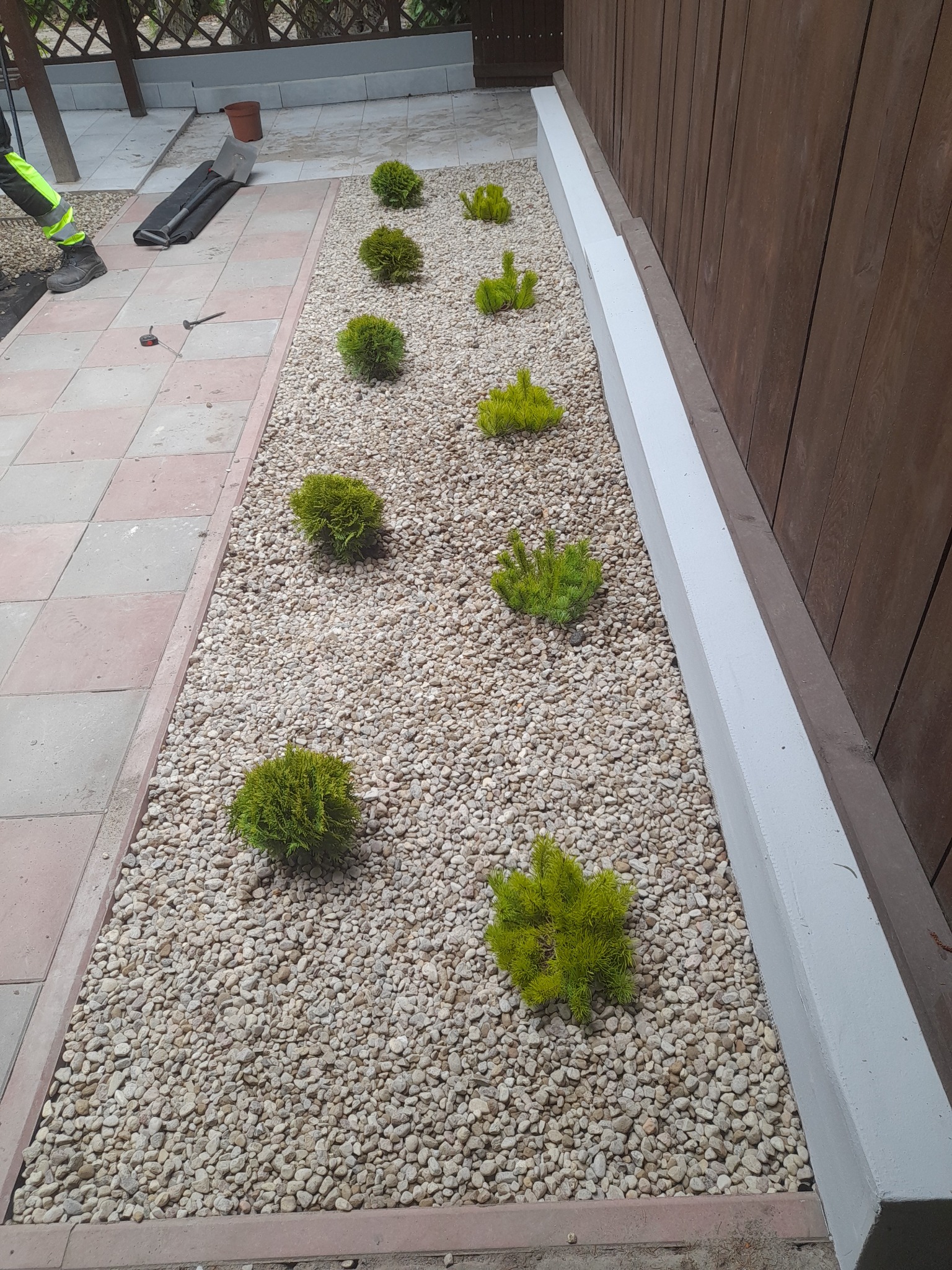 Aranżacja ogrodu z kulistymi krzewami iglastymi i drobnym białym grysem, widoczne narzędzia ogrodnicze i fragment stroju roboczego ogrodnika.