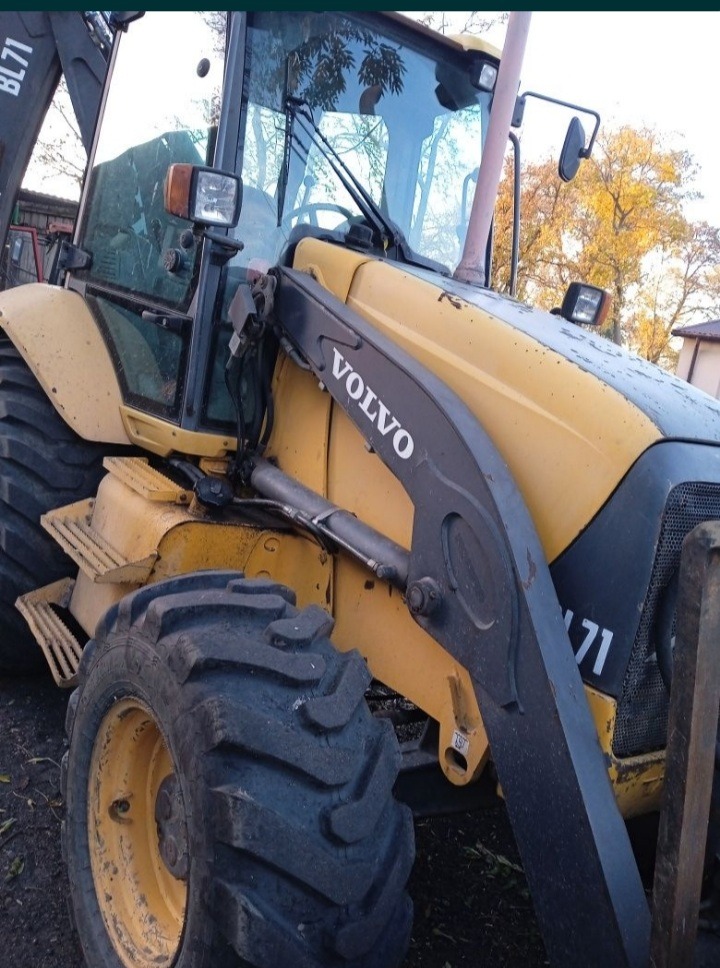 Żółta ładowarka VOLVO BL71 na ziemi, widok z bliska na oponę i ramię ładowarki, drzewo z żółtymi liśćmi w tle.