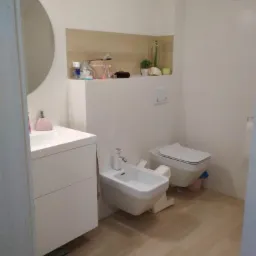 Nowoczesna łazienka z białymi sanitariatami: umywalka z szafką, bidet i toaleta podwieszana, wykończona płytkami imitującymi drewno na podłodze i częściowo na ścianie, z dekoracyjną półką nad toaletą.