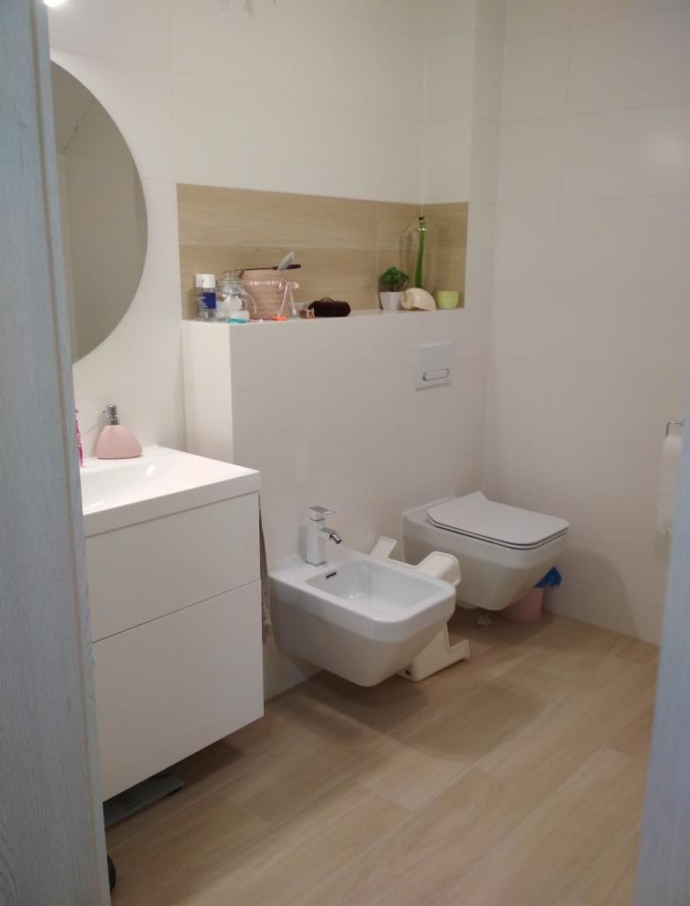 Nowoczesna łazienka z białymi sanitariatami: umywalka z szafką, bidet i toaleta podwieszana, wykończona płytkami imitującymi drewno na podłodze i częściowo na ścianie, z dekoracyjną półką nad toaletą.