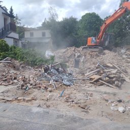 Mila&Trans Sp. z o.o - Pomarańczowa koparka Hitachi na gruzowisku po rozbiórce budynku, widoczny fragment budynku w tle, pracownik w pomarańczowej koszulce nadzoruje załadunek gruzu na ciężarówkę.