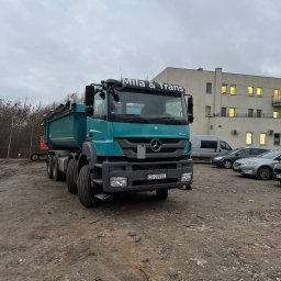 Mila&Trans Sp. z o.o - Turkusowa wywrotka Mercedes-Benz z napisem 'MILA & TRANS' na tle placu budowy z gruzem i zaparkowanymi samochodami, widoczny budynek z oświetlonymi oknami.