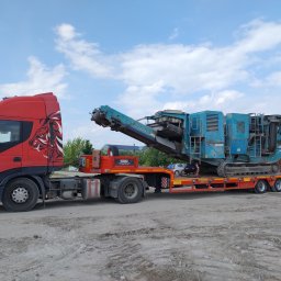 Mila&Trans Sp. z o.o - Czerwona ciężarówka z grafiką lwa transportuje na niskopodwoziowej platformie niebieską, gąsienicową kruszarkę do kamienia; operator w kucki sprawdza mocowanie ładunku.