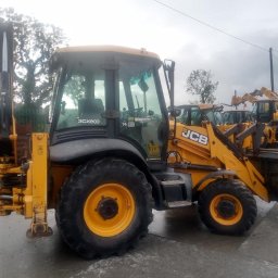 Mila&Trans Sp. z o.o - Żółta koparko-ładowarka JCB 3CX ECO na placu z innymi maszynami, widoczne opony z żółtymi felgami i metalowa krata w tle.