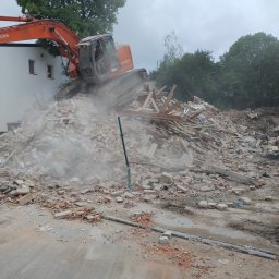 Mila&Trans Sp. z o.o - Pomarańczowa koparka Hitachi na szczycie sterty gruzu i cegieł podczas rozbiórki budynku, widoczny operator w kabinie, w tle biały budynek i drzewa.