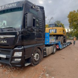 Mila&Trans Sp. z o.o - Czarny ciągnik siodłowy MAN z naczepą niskopodwoziową przewożący żółtą koparkę kołową CAT, widoczny pracownik w odblaskowej kamizelce, teren budowy w tle.