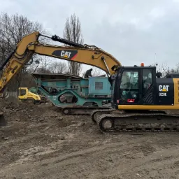 Żółto-czarny koparka CAT 323E z łyżką i przesiewacz Pegson na placu budowy z ziemią, w tle żółty walec drogowym, pochmurne niebo.