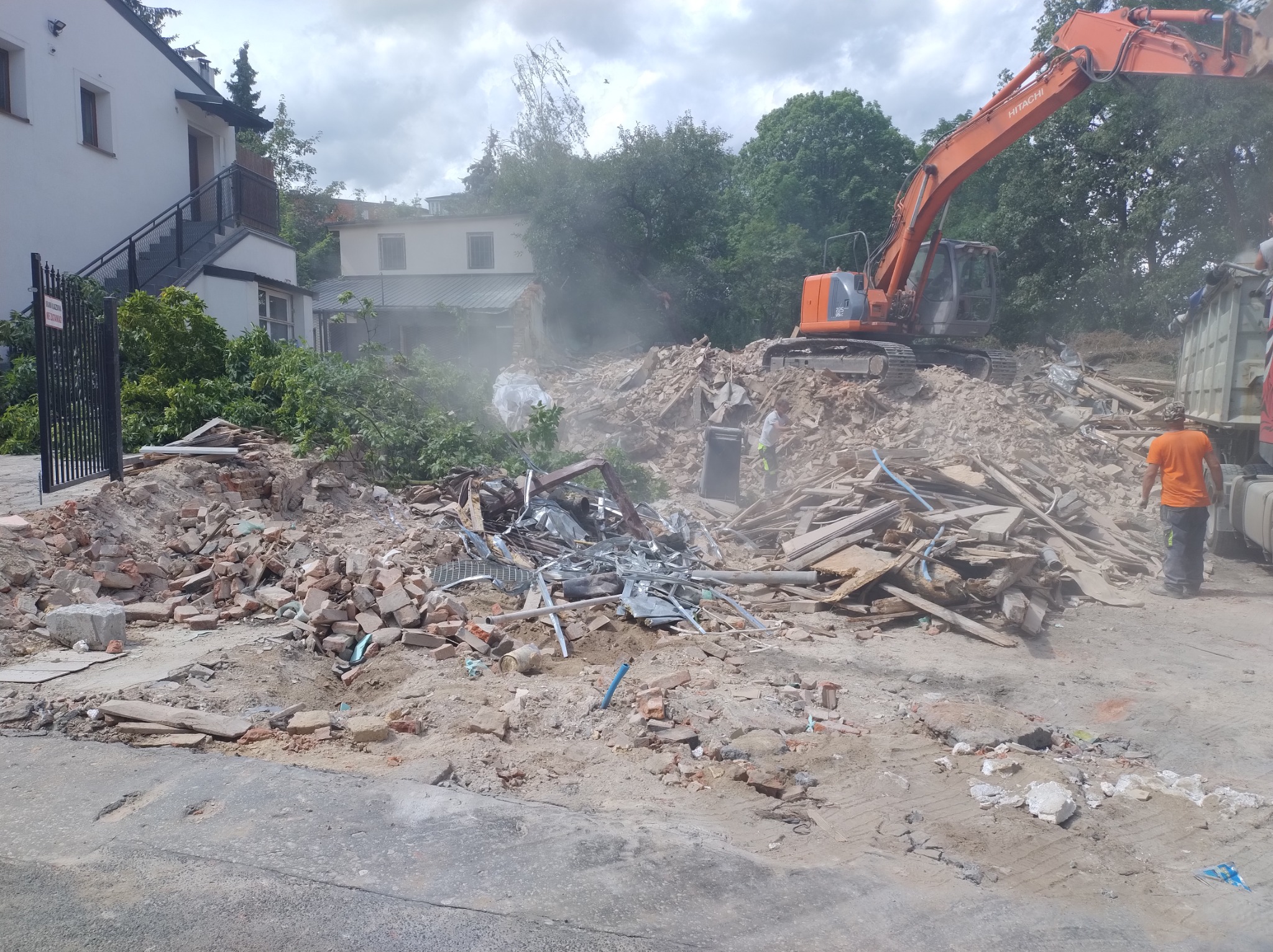 Pomarańczowa koparka Hitachi na gruzowisku po rozbiórce budynku, widoczny fragment budynku w tle, pracownik w pomarańczowej koszulce nadzoruje załadunek gruzu na ciężarówkę.