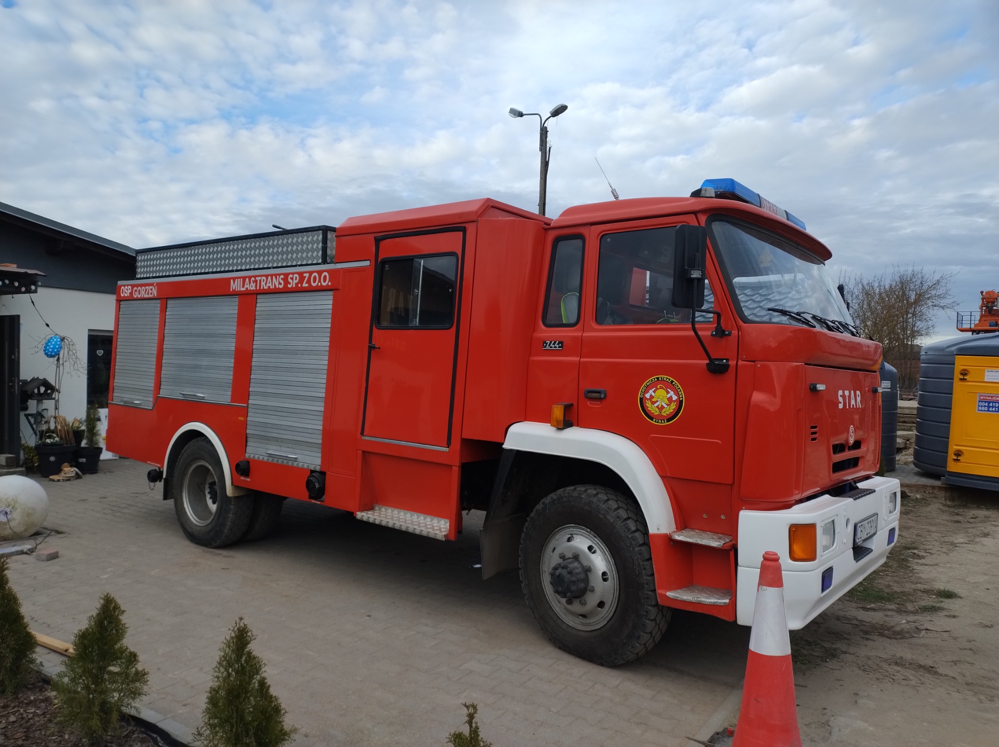 Czerwony wóz strażacki marki Star z napisem 'MILA&TRANS SP. Z O.O.' zaparkowany na kostce brukowej, z widocznym stożkiem ostrzegawczym i elementami krajobrazu w tle.