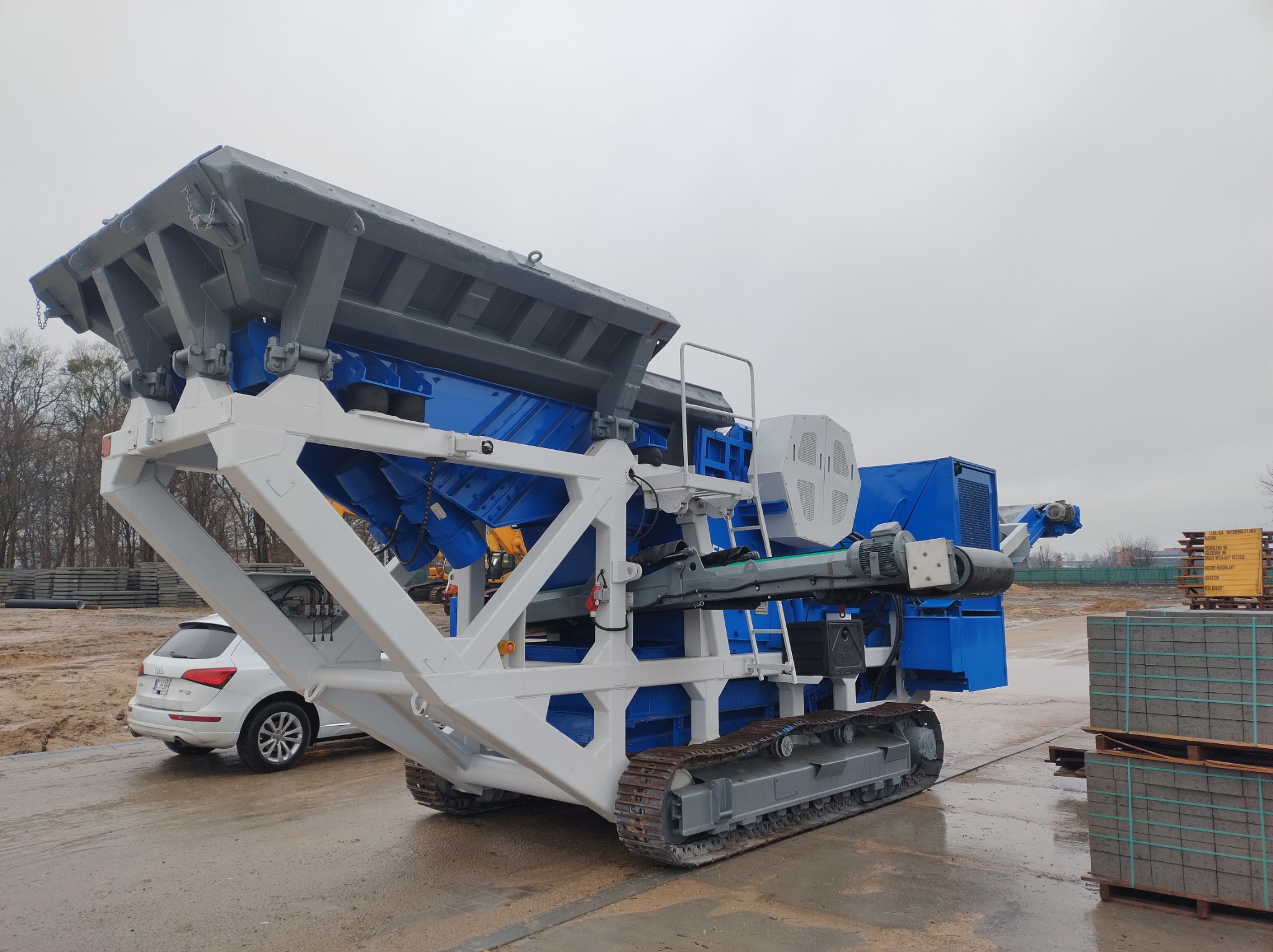 Mobilna kruszarka na gąsienicach, pomalowana na biało i niebiesko, stoi na betonowym placu w pochmurny dzień. W tle zaparkowany biały samochód osobowy i palety z kostką brukową.