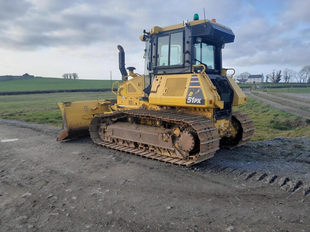 Żółty spychacz gąsienicowy D51PX stoi na ubitej, błotnistej drodze polnej, w tle zielone wzgórza i zabudowania gospodarcze pod pochmurnym niebem.