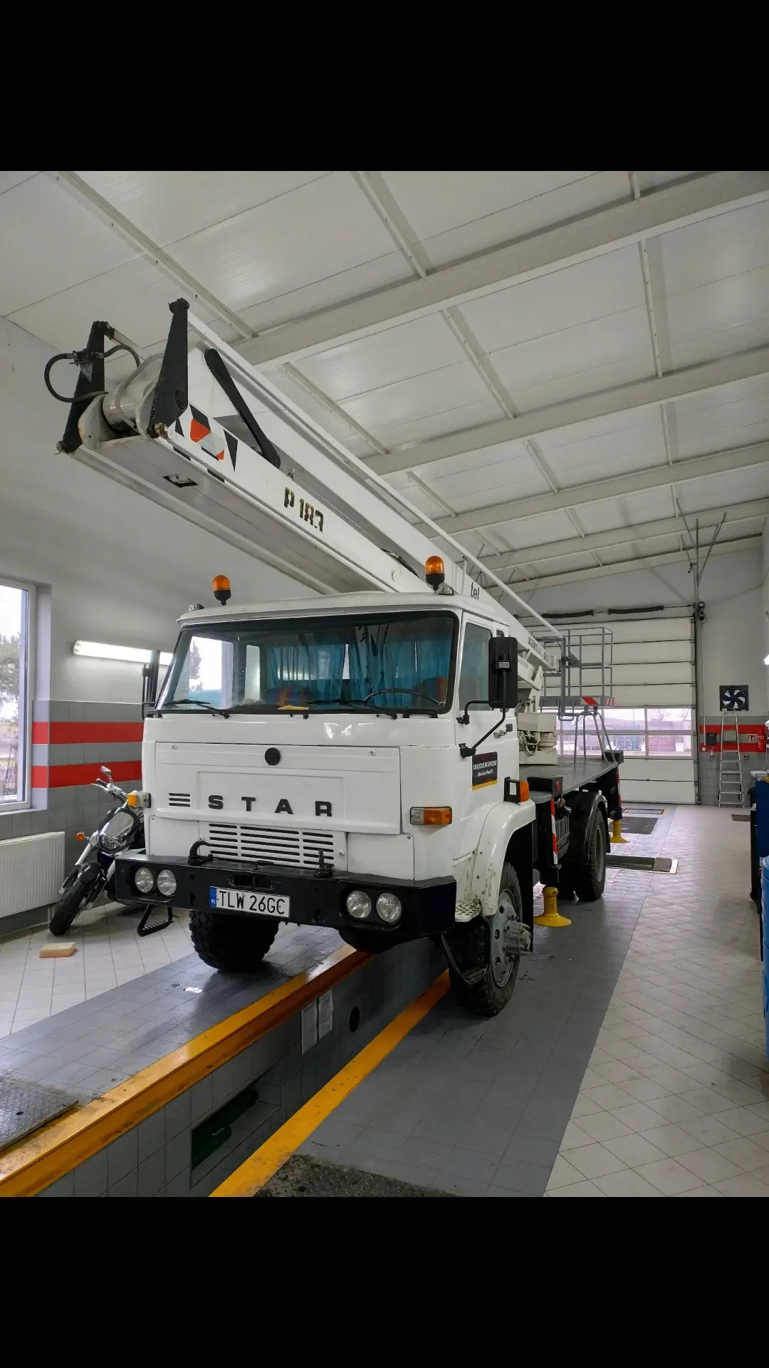 Biały samochód ciężarowy STAR z podnośnikiem koszowym P183 w warsztacie, widoczny motocykl w tle, tablica rejestracyjna TLW 26GC.