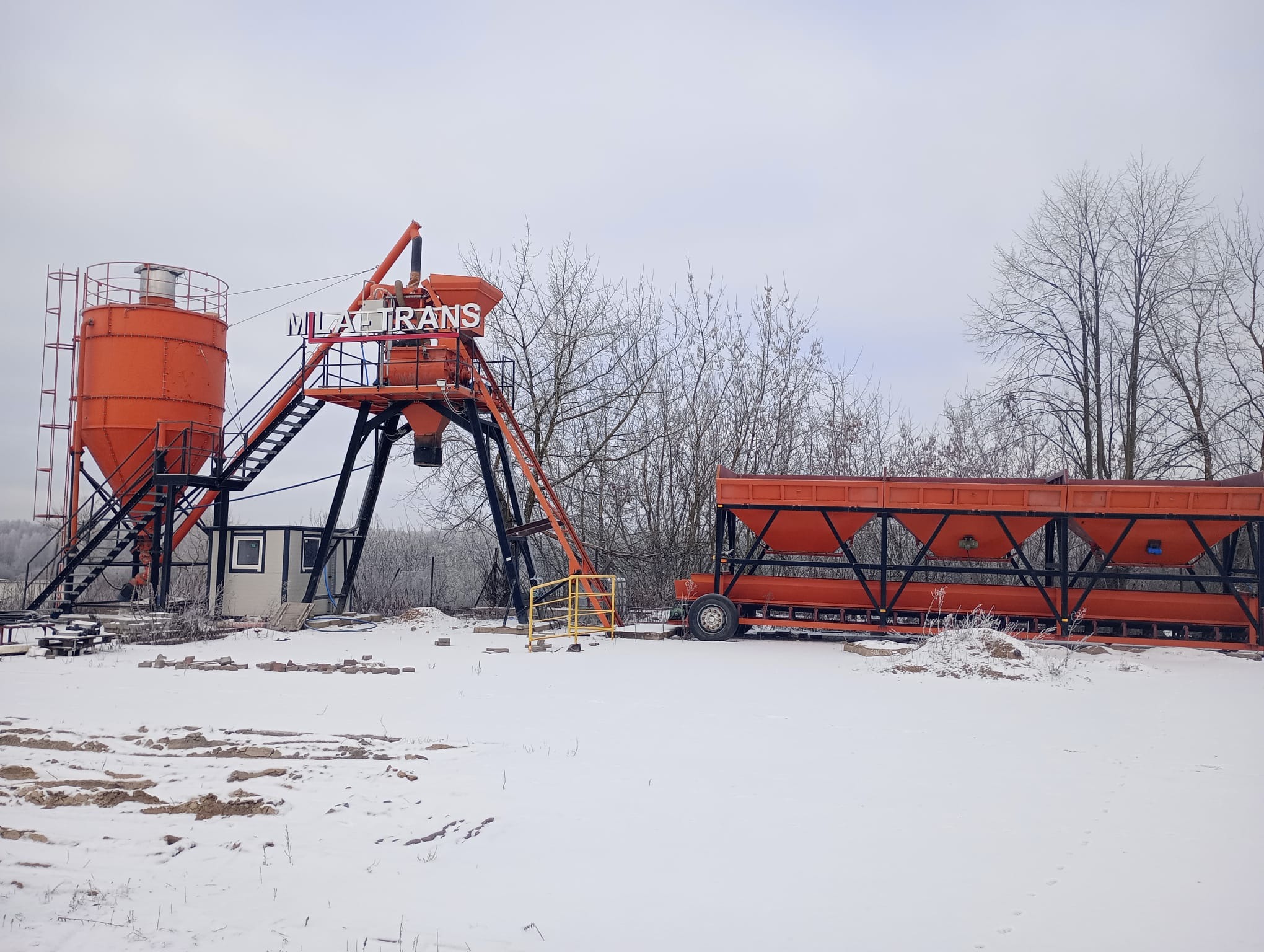 Pomarańczowa betoniarnia z napisem 'MILA TRANS' na tle zimowego krajobrazu, śnieg na ziemi, widoczne drzewa bez liści.