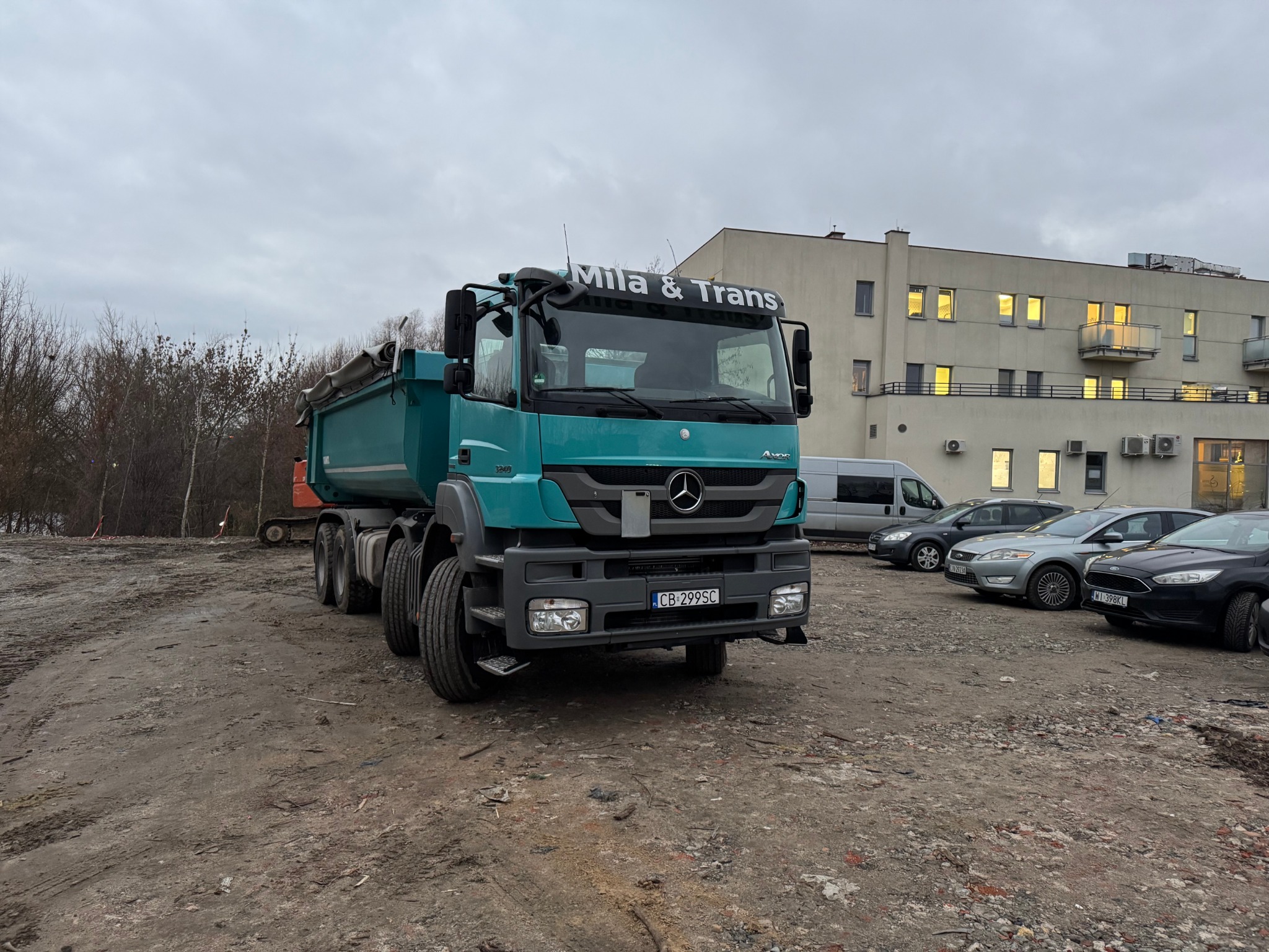 Turkusowa wywrotka Mercedes-Benz z napisem 'MILA & TRANS' na tle placu budowy z gruzem i zaparkowanymi samochodami, widoczny budynek z oświetlonymi oknami.