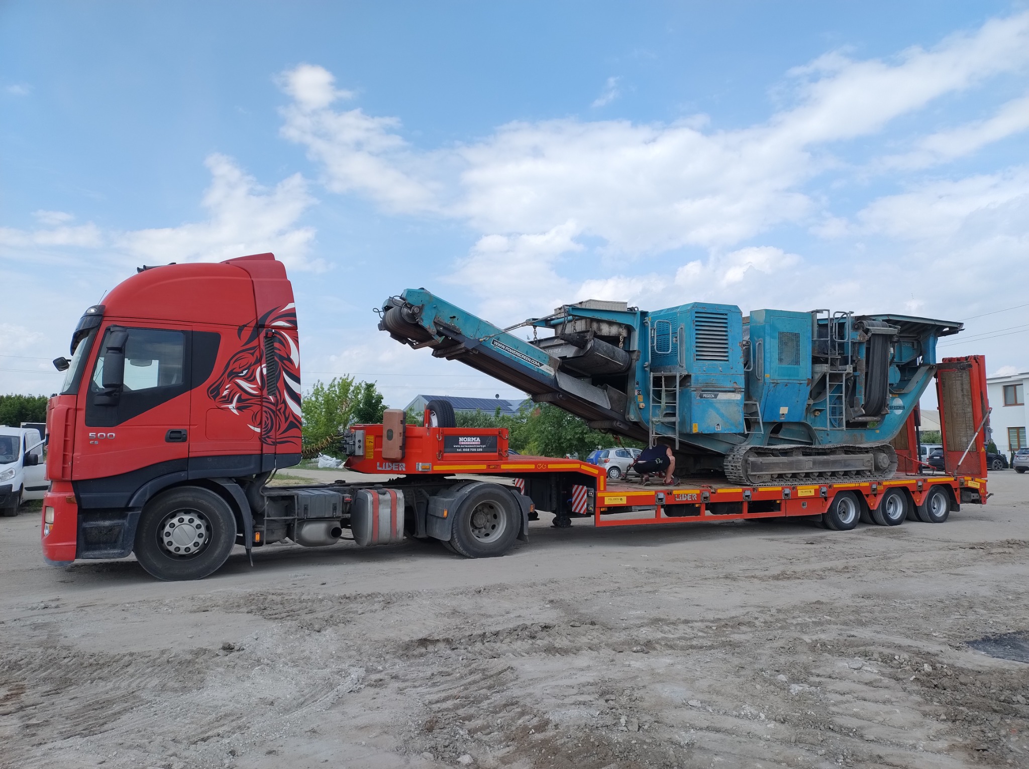 Czerwona ciężarówka z grafiką lwa transportuje na niskopodwoziowej platformie niebieską, gąsienicową kruszarkę do kamienia; operator w kucki sprawdza mocowanie ładunku.