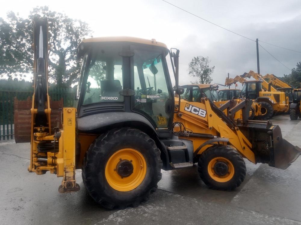 Żółta koparko-ładowarka JCB 3CX ECO na placu z innymi maszynami, widoczne opony z żółtymi felgami i metalowa krata w tle.