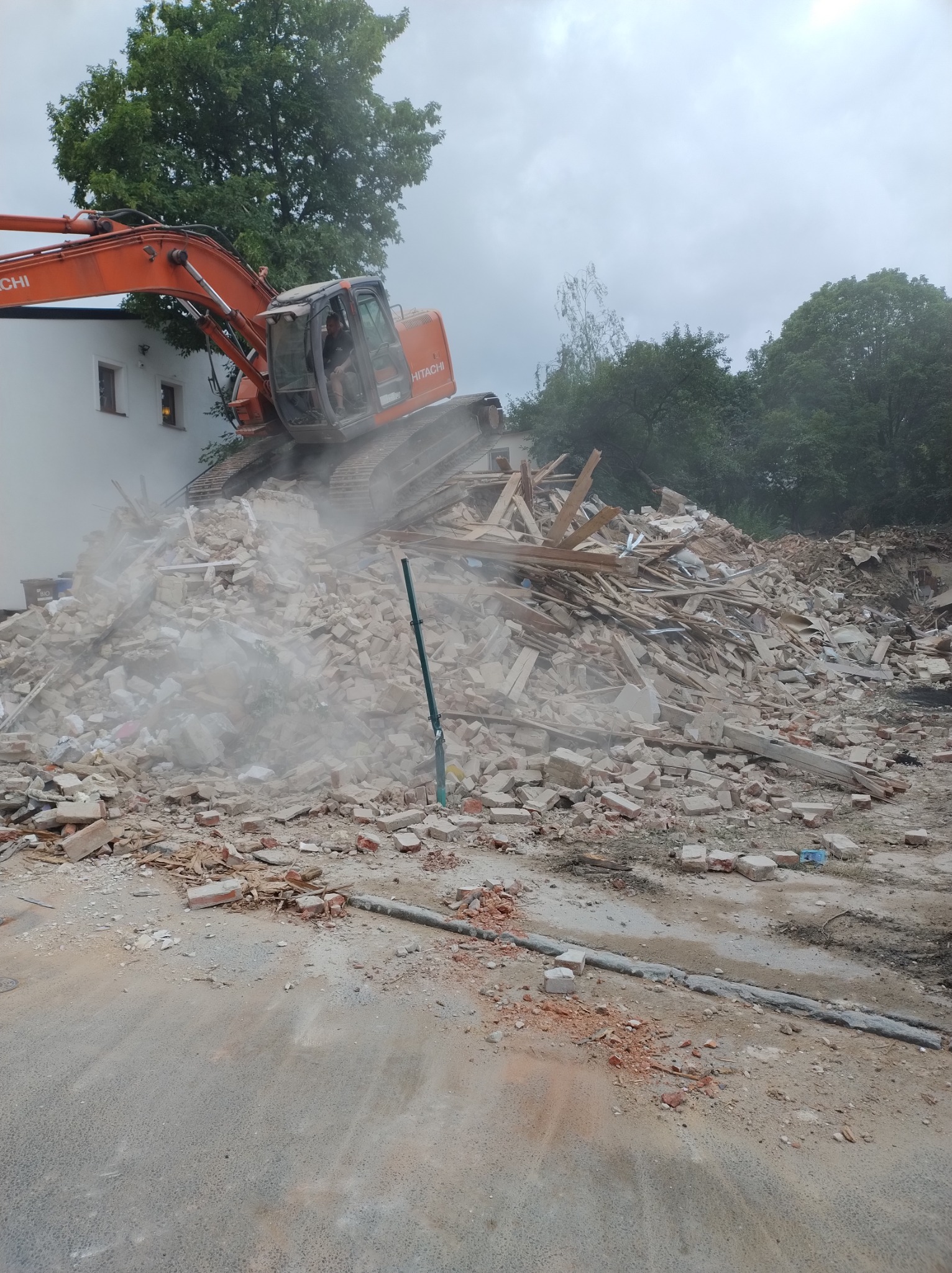 Pomarańczowa koparka Hitachi na szczycie sterty gruzu i cegieł podczas rozbiórki budynku, widoczny operator w kabinie, w tle biały budynek i drzewa.