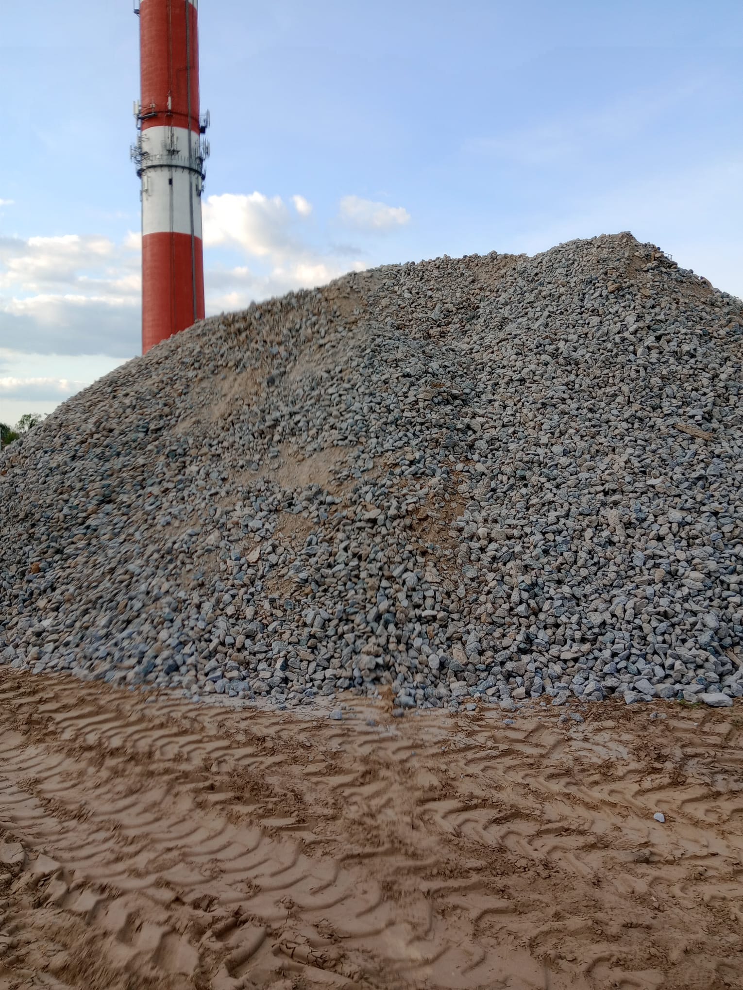 Góra szarego kruszywa na ziemi z wyraźnymi śladami opon, w tle wysoki komin z czerwono-białymi pasami i częściowo zachmurzone niebo.