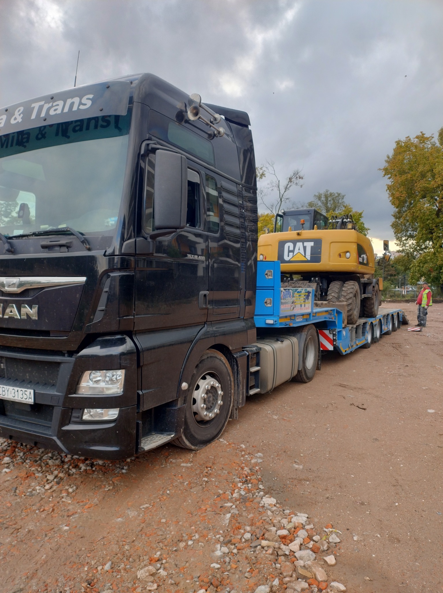 Czarny ciągnik siodłowy MAN z naczepą niskopodwoziową przewożący żółtą koparkę kołową CAT, widoczny pracownik w odblaskowej kamizelce, teren budowy w tle.
