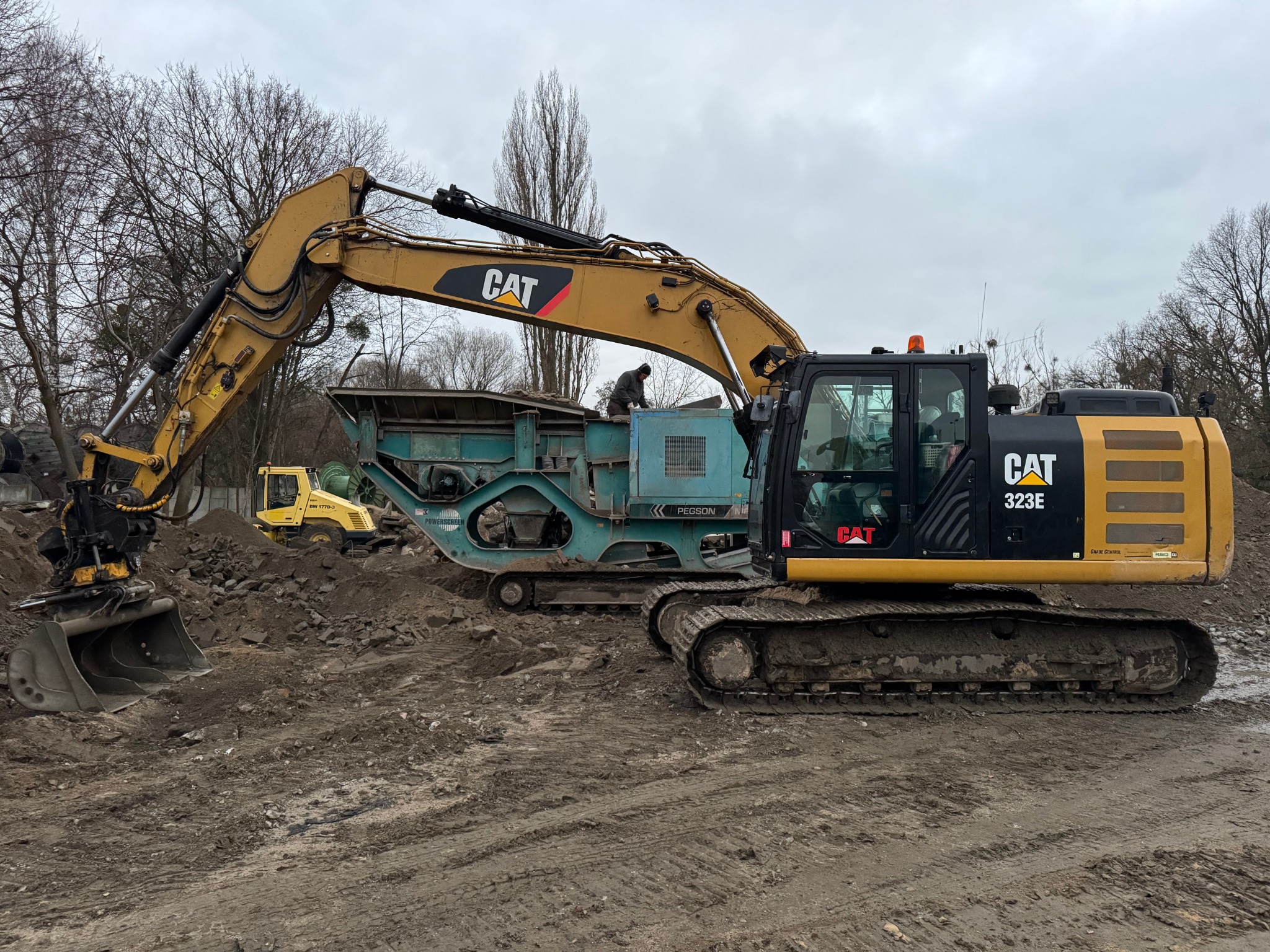 Żółto-czarny koparka CAT 323E z łyżką i przesiewacz Pegson na placu budowy z ziemią, w tle żółty walec drogowym, pochmurne niebo.