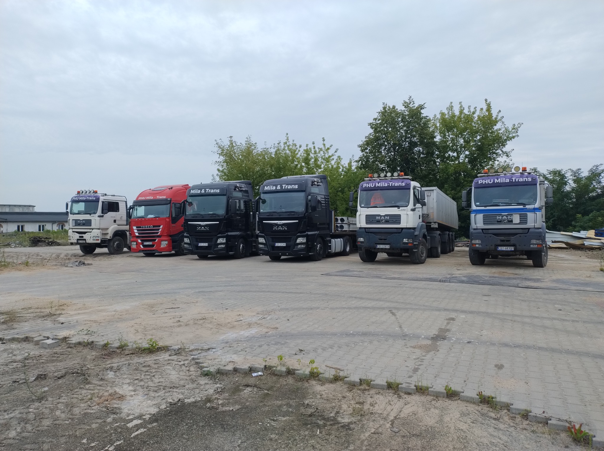 Flota pojazdów ciężarowych MAN i Iveco firmy transportowej PHU Mila-Trans, zaparkowana na placu z kostki betonowej w pochmurny dzień.