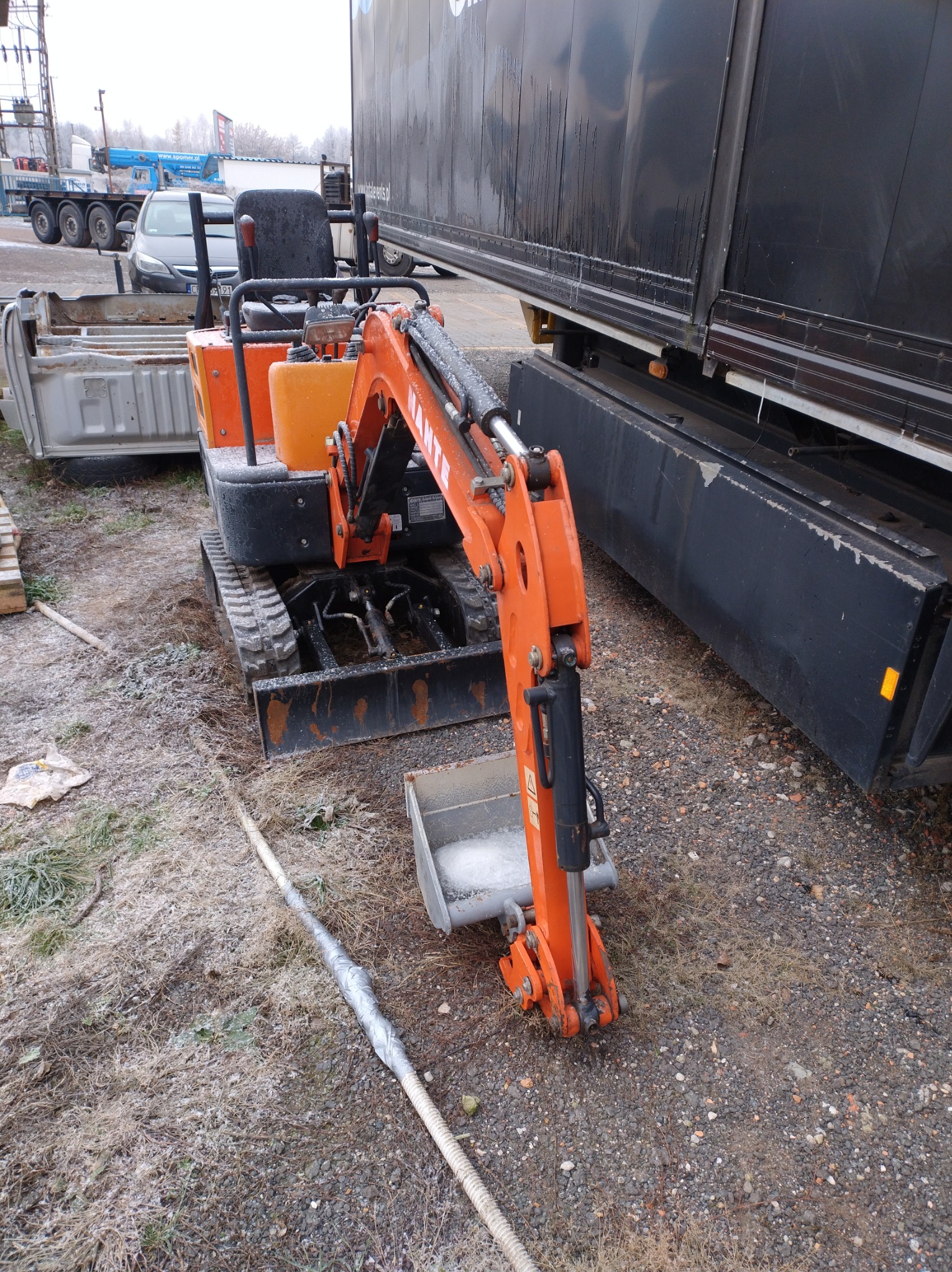 Pomarańczowa mini koparka Hante na placu budowy, obok zaparkowana ciężarówka z czarną plandeką, ziemia pokryta szronem.