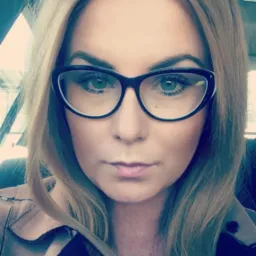 Portret kobiety w okularach z czarną oprawką, o jasnych włosach i zielonych oczach, w samochodzie.