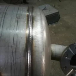 Cylindryczny zbiornik ze stali nierdzewnej z widocznymi spawami i przyłączem kołnierzowym, w tle widoczne elementy wyposażenia warsztatowego.